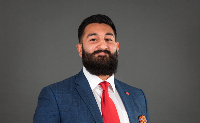 Baljinder poses wearing a blue suit. He wears a red necktie with an orange pocket square.