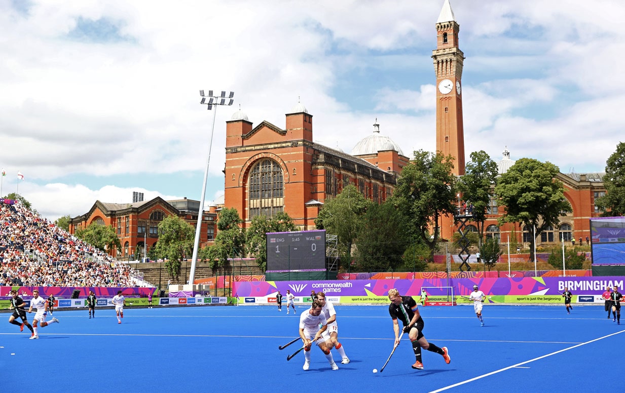 The men's GB hockey team in action on a blue hockey court with the Aston Webb building and Old Joe in the background