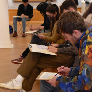 students sit on stools in the Barber art gallery, quietly sketching on a clipboard resting on their laps.
