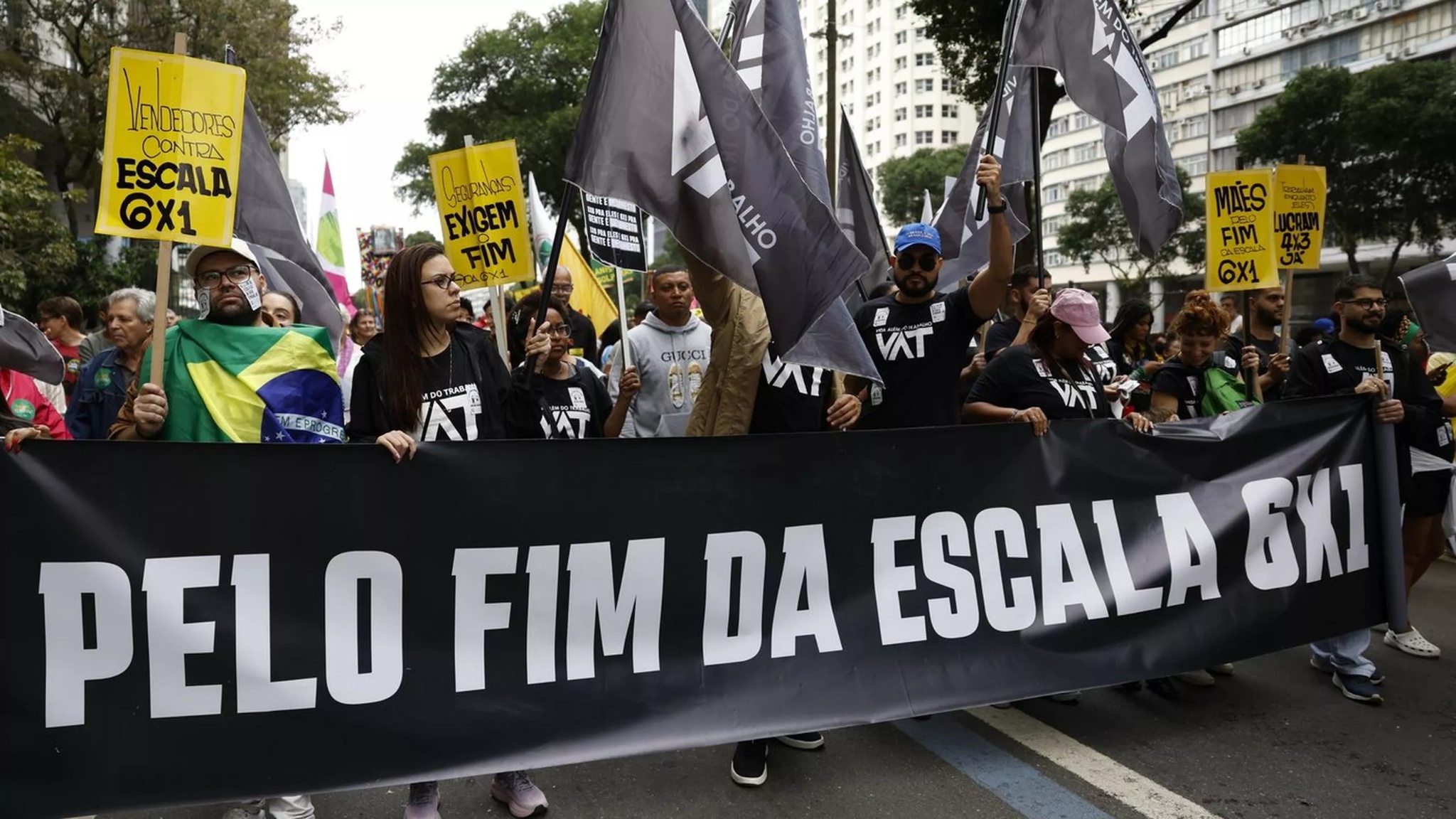 People march along a city street carrying flags and protest signs behind a large black banner