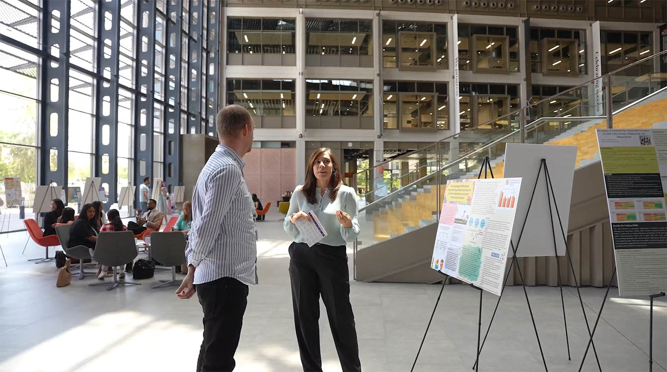 Two people standing in front of a poster with research on it. They are standing in a open building space 