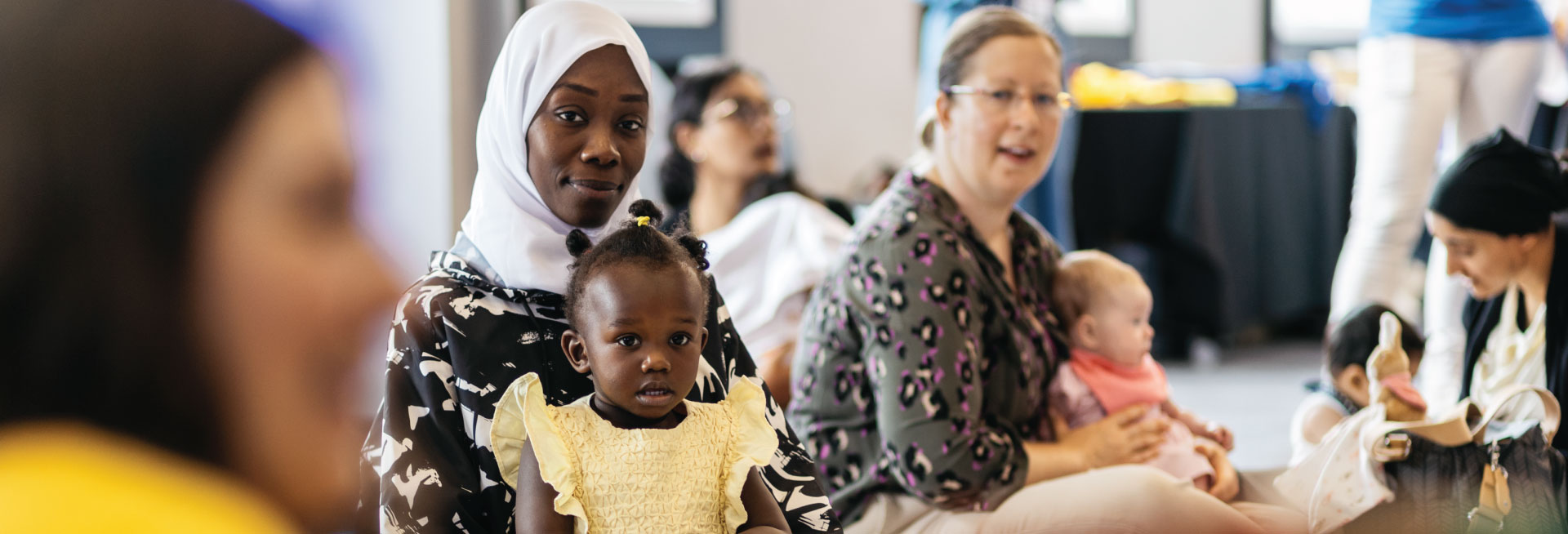 A room of mothers and babies listen to a presenter reading a book.