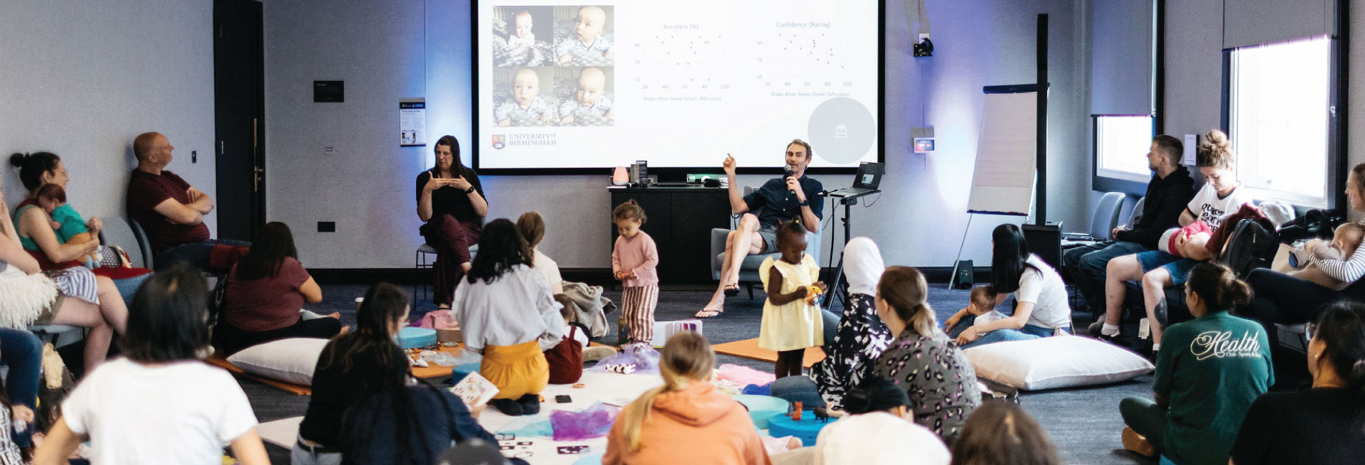 A room full of parents and carers and their children sit and listen to a presenter in front of a large screen.