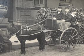Black and white image of three children sitting in a cart behind a pony