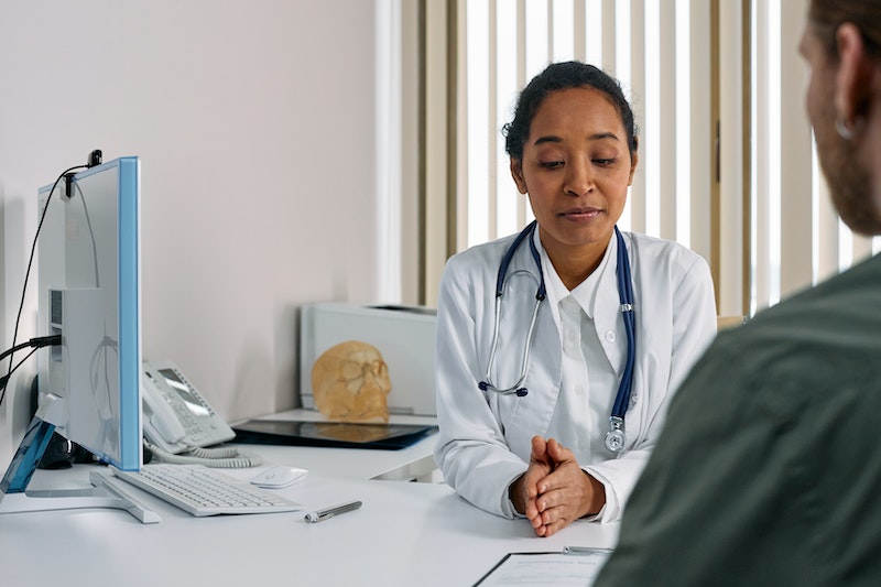 Black female doctor in consultation room sat at a desk which has computer, printer and plastic model of a human skull talking to a patient