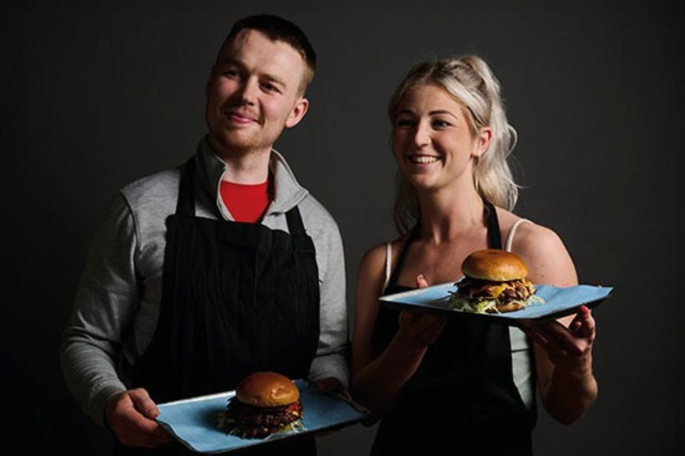Two people stand wearing black aprons and holding burgers on serving trays.