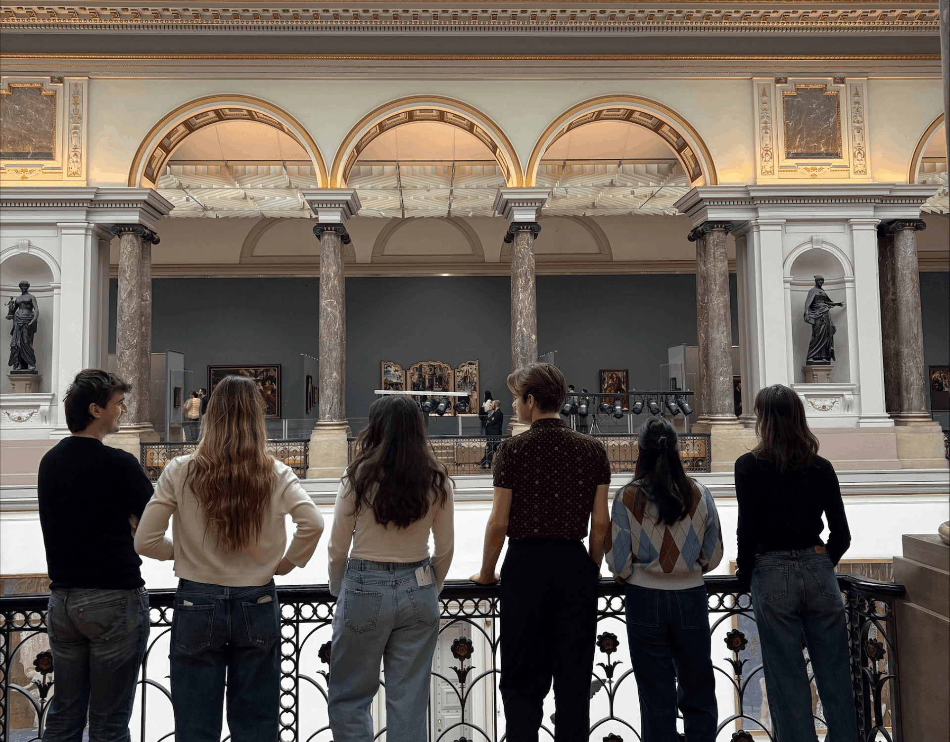 A group of six students stand on a gallery balcony looking out ahead of them at the art works 