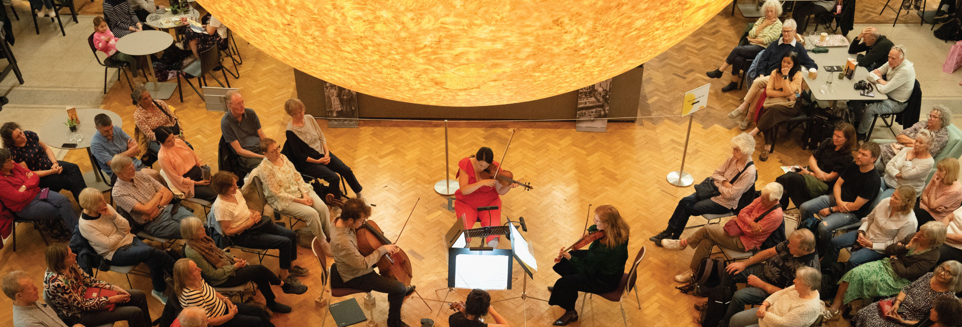 String musicians perform under an illuminated sculpture of the Sun to an audience in The Exchange.