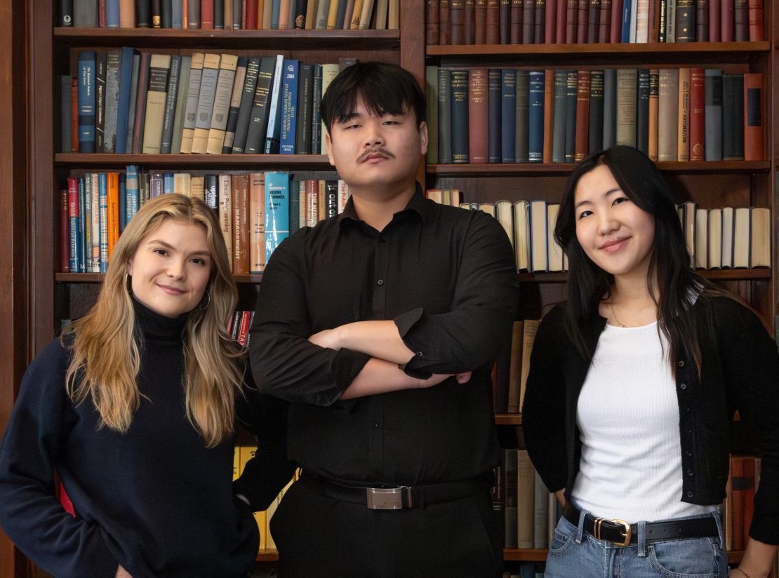 Three students stand in front of a bookshelf, smiling at the camera. 