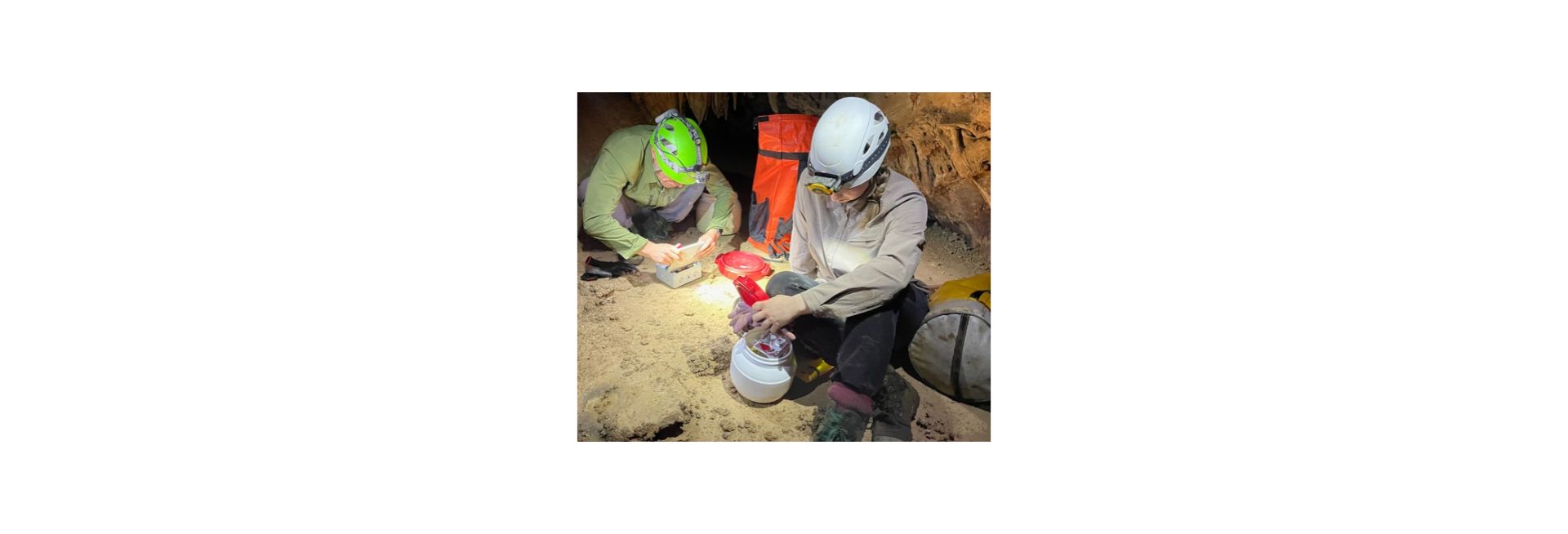 Two researchers wearing hard hats conduct fieldwork in a cave environment.