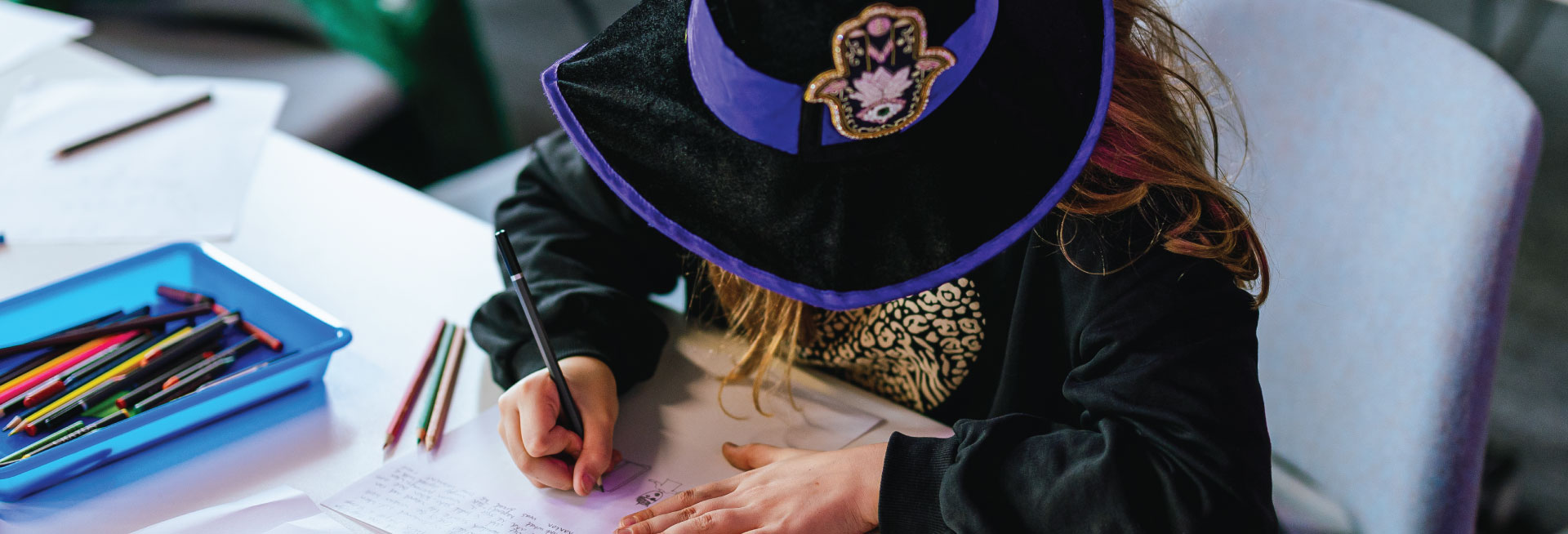 A child dressed as a witch does some writing with colouring pencils.