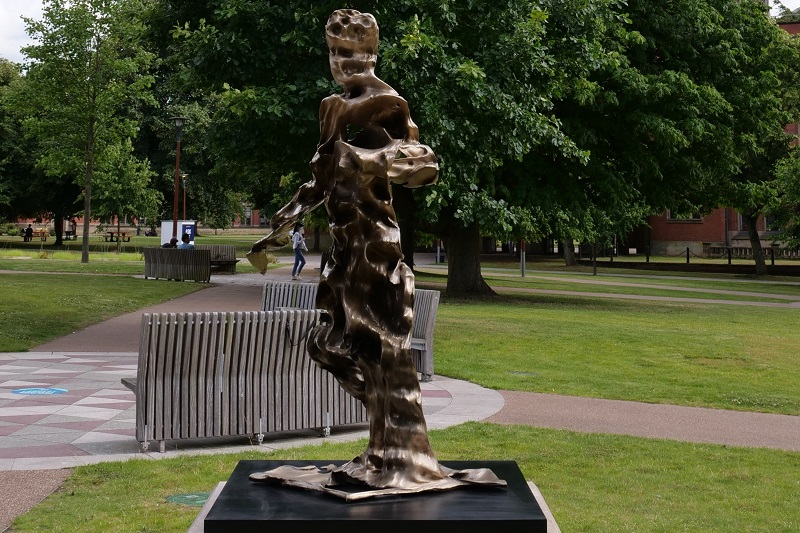Bronze sculpture called Father Sky by artist Zachary Eastwood-Bloom installed on the University of Birmingham Edgbaston campus on a black plinth. Picture is from an angle showing sculpture's face and human shape with benches and trees in background.