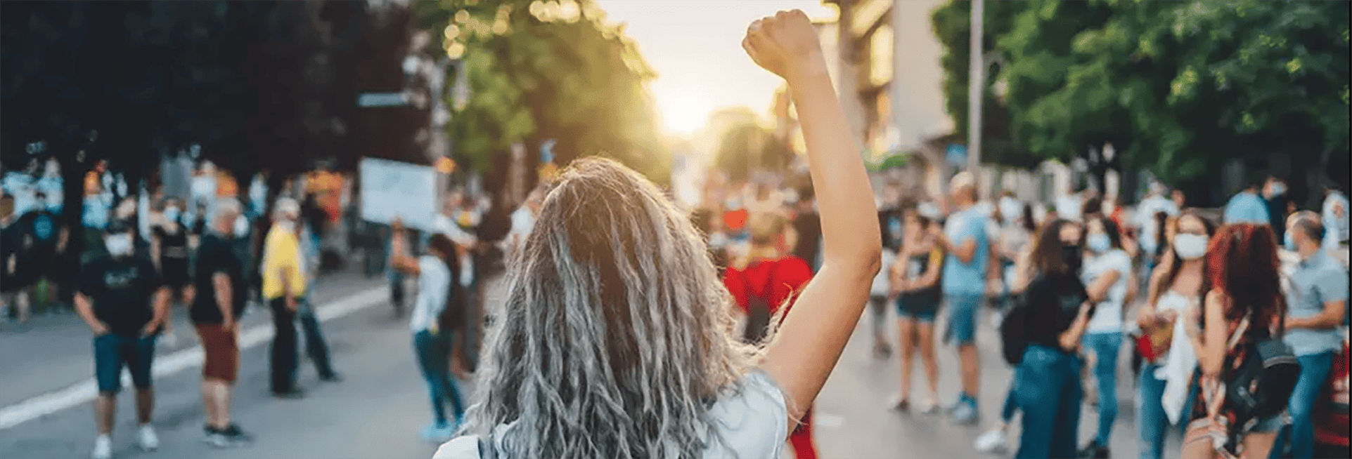 Back of the head of a young woman at a protest with her fist in the air and a crowd of protesters in front of her.