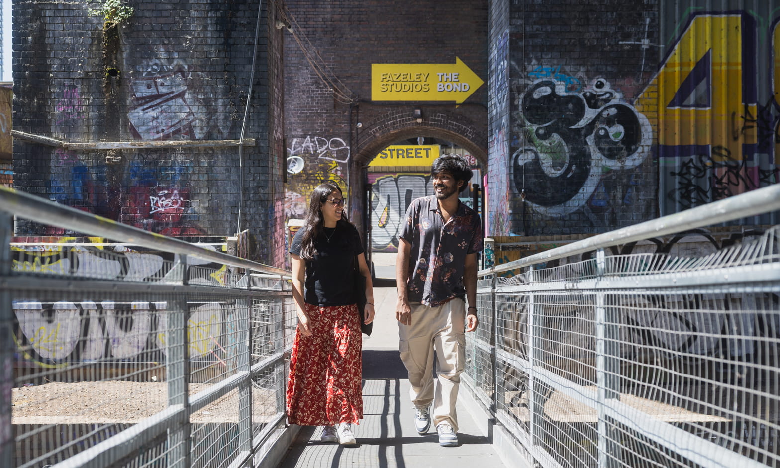 Two smiling students walk across a metal bridge in Digbeth. Behind them, there is a graffiti-covered wall