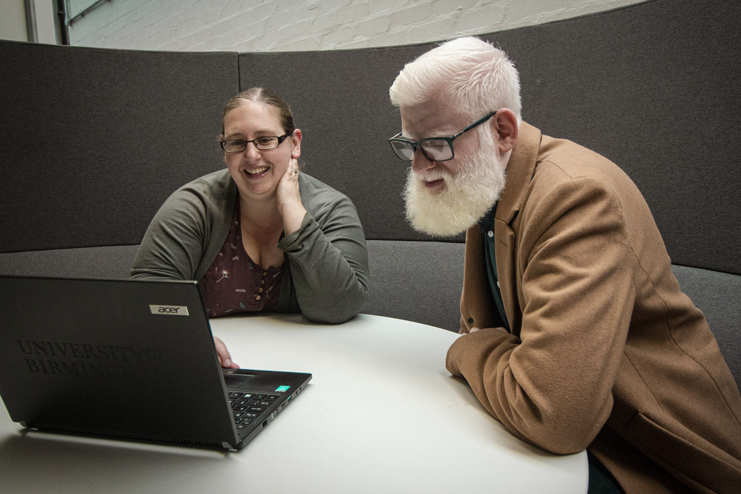 A man and woman, both wearing glasses, look at a laptop