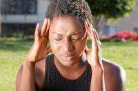 Young black woman with black sports top sat with hands raised to press her fingers to her temples and an expression of pain on her face. Background is out of focus image of a park scene with trees and grass