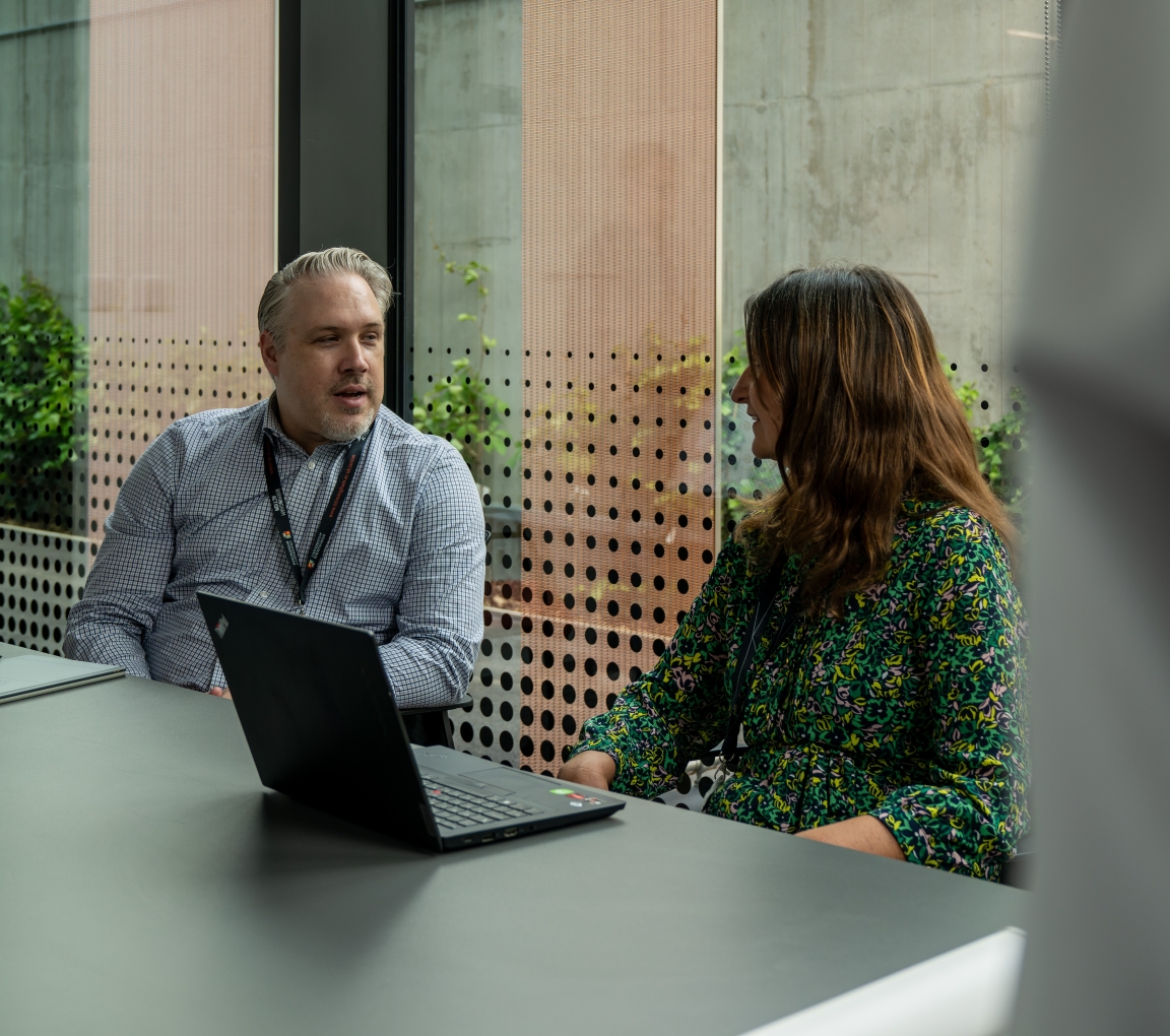 Two people sat at a desk in conversation with a laptop.