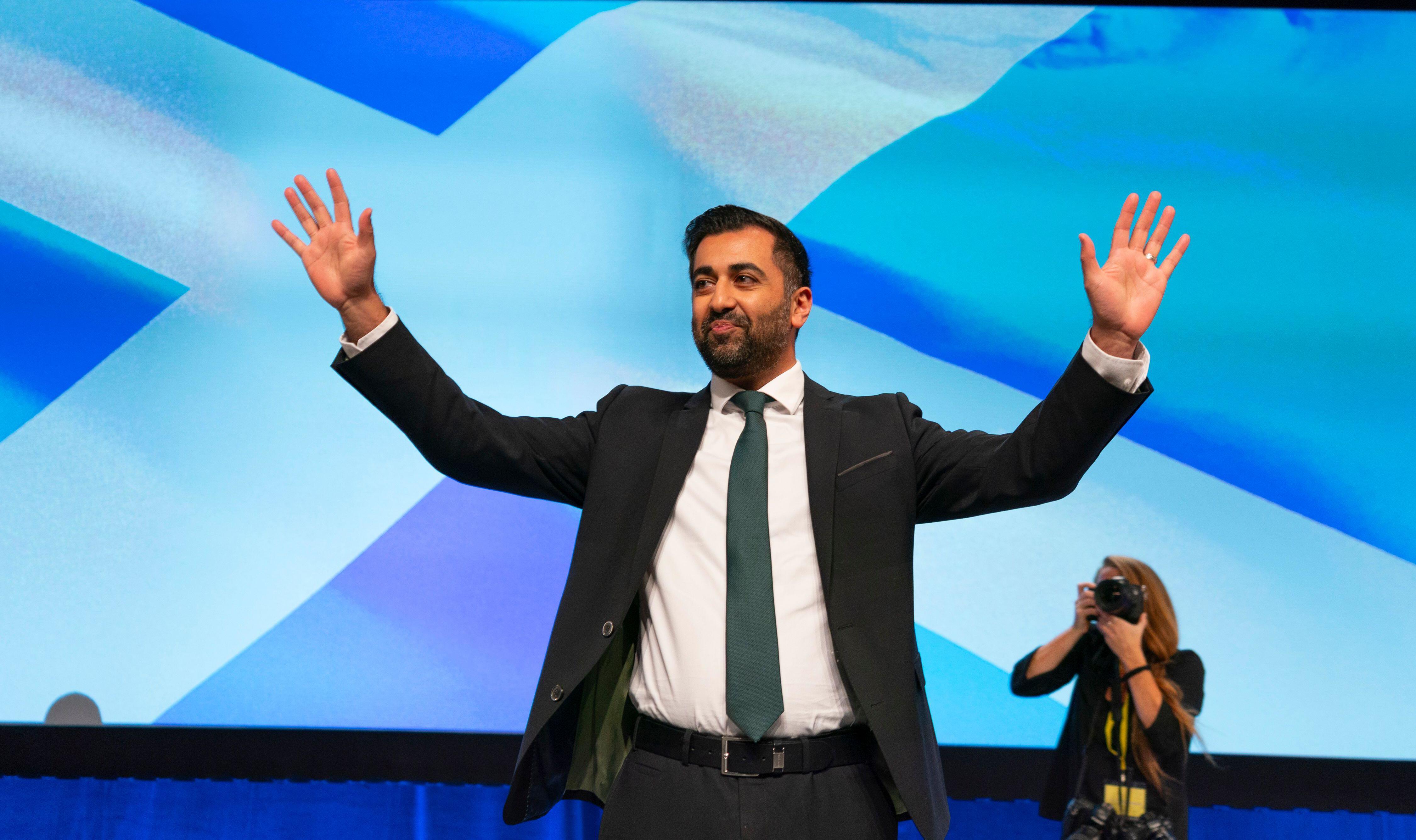 Humza Yousaf being photographed waving at a crowd with the Scottish flag in the background.