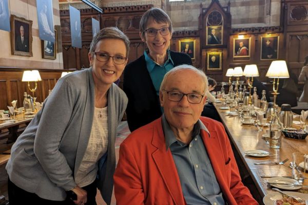 Three people (two women and one man, who is seated) smiling at the camera in a dining hall.