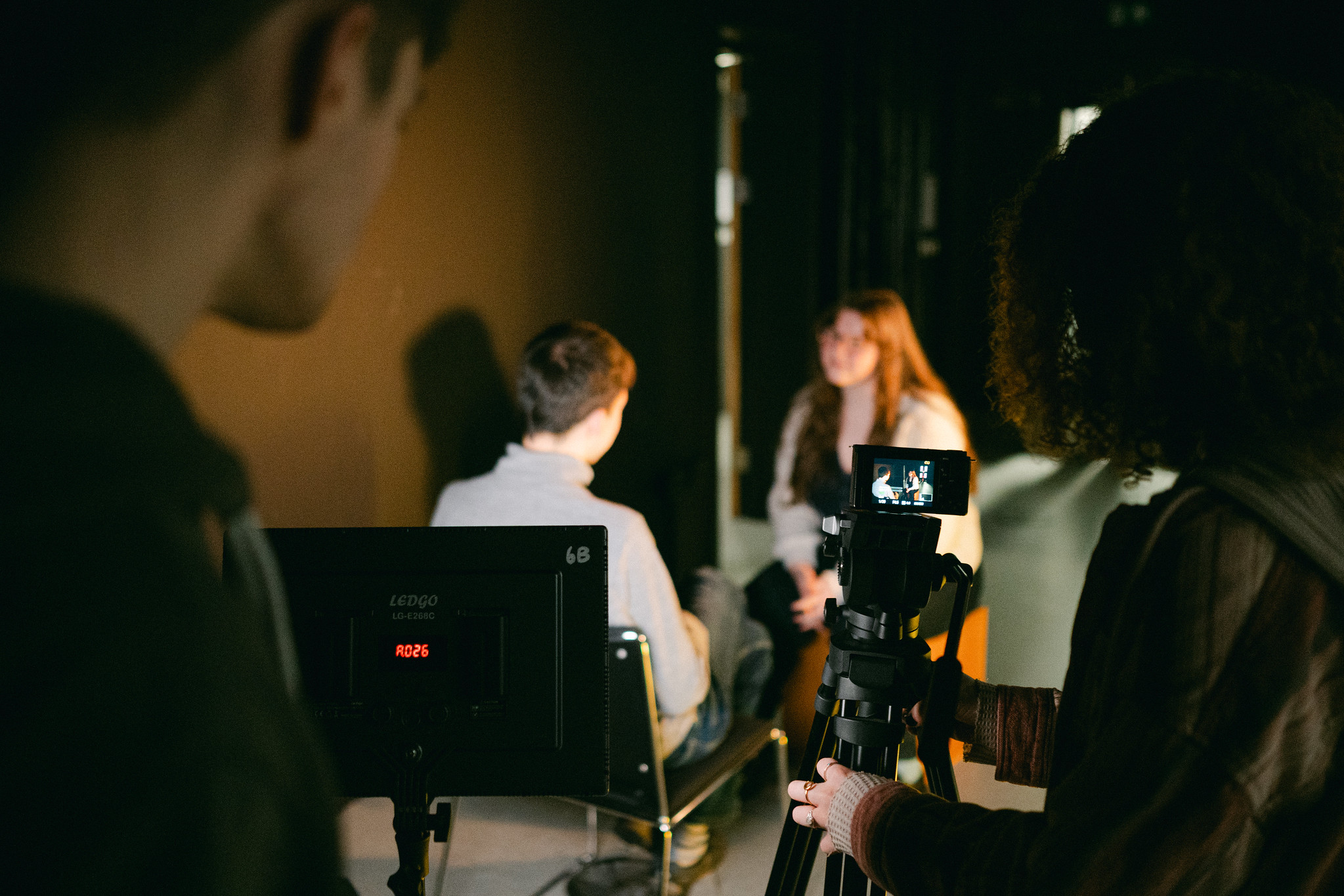 Two students standing behind a camera and lighting rig filming an interview in a studio space.