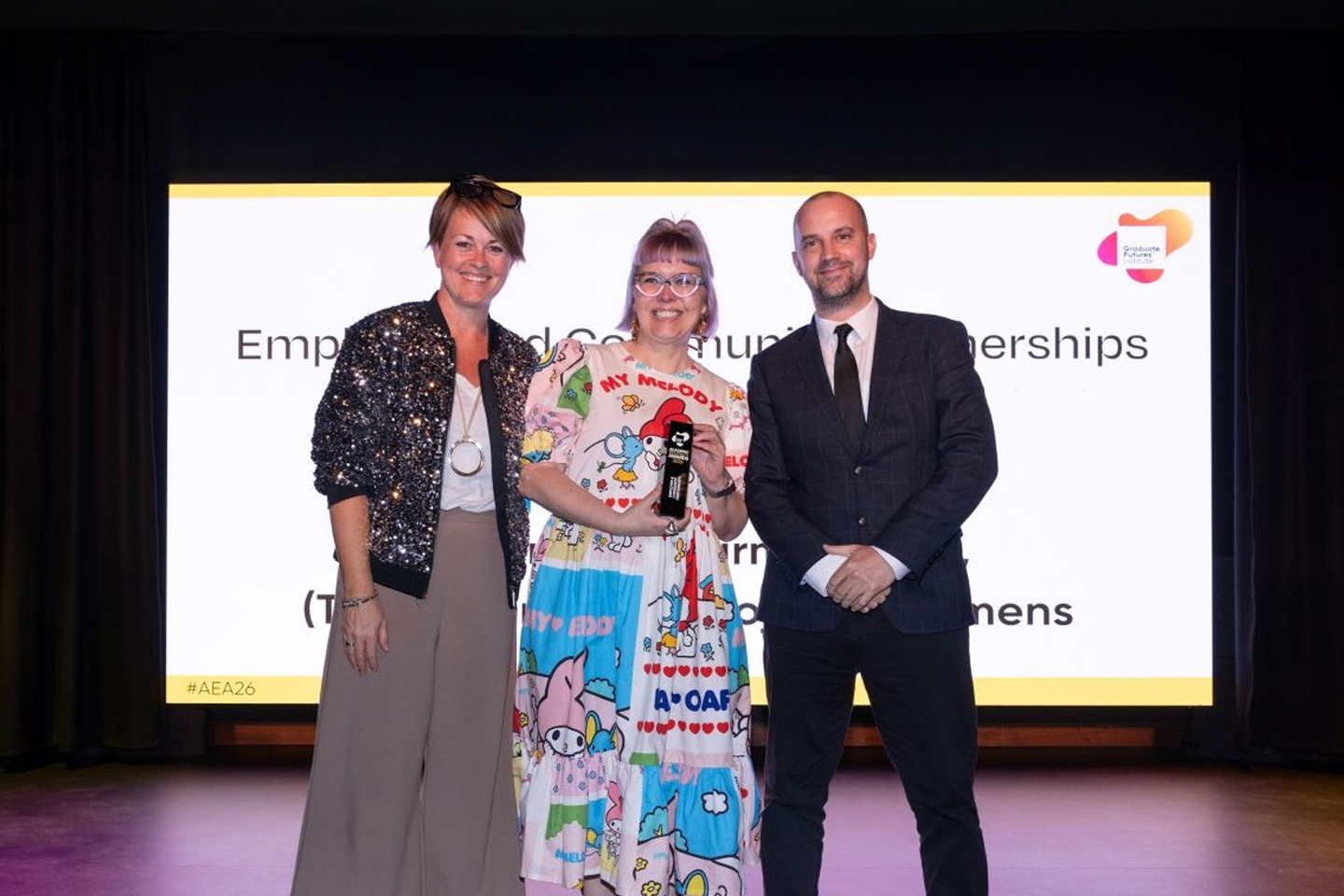 Dr Sarah Hall standing on stage holding an award flanked by Dr Brooke Storer-Church and Bob Lee