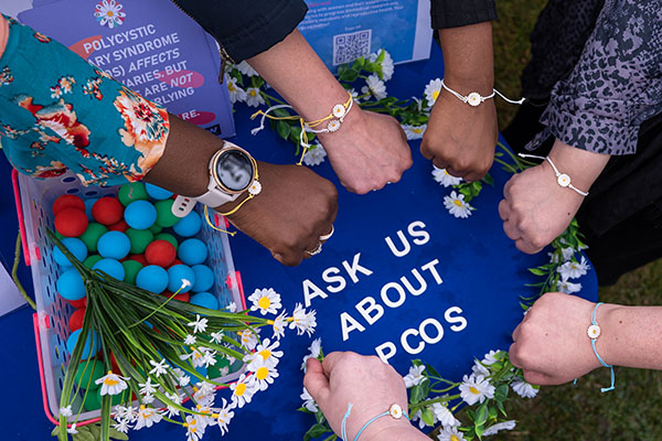 Lots of hands with daisy bracelets around their wrists in front of wording that says 'Ask us about PCOS'