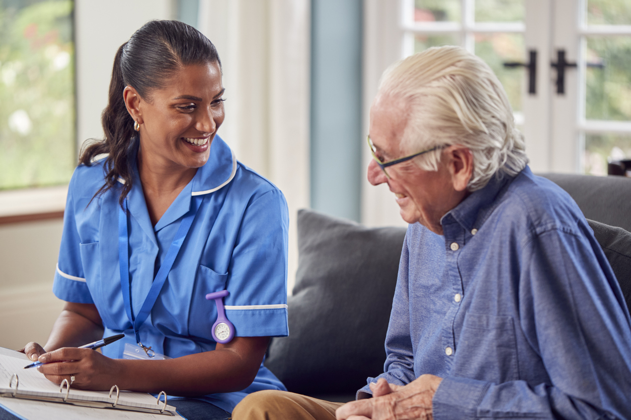 A home visit by a nurse (left) with an elderly male patient (right). The nurse and patient are smiling and talking to each other.