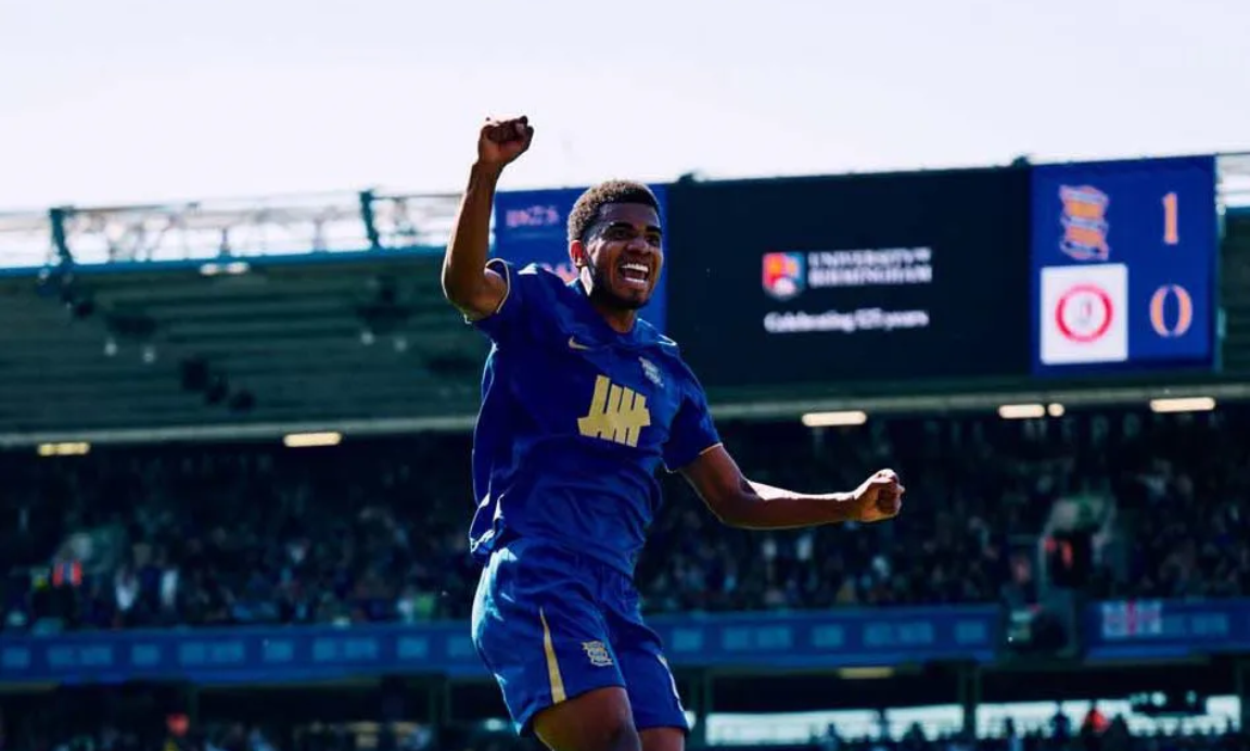 A man in football kit is jumping with his arm raised, in a football stadium