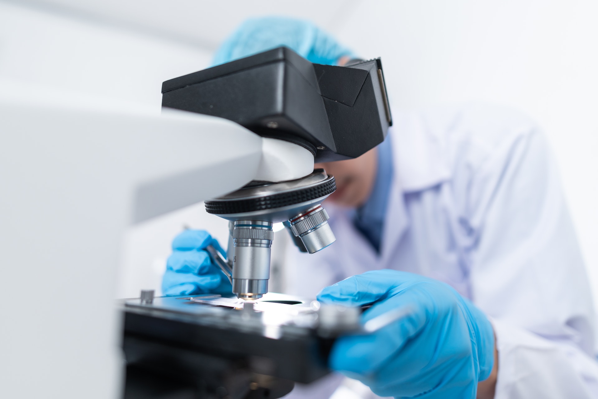 Masked scientist, wearing protective glasses and white lab coat, looking into a microscope.