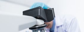 Microscope in the foreground with female biomedical scientist out of focus examining a sample on a plate 