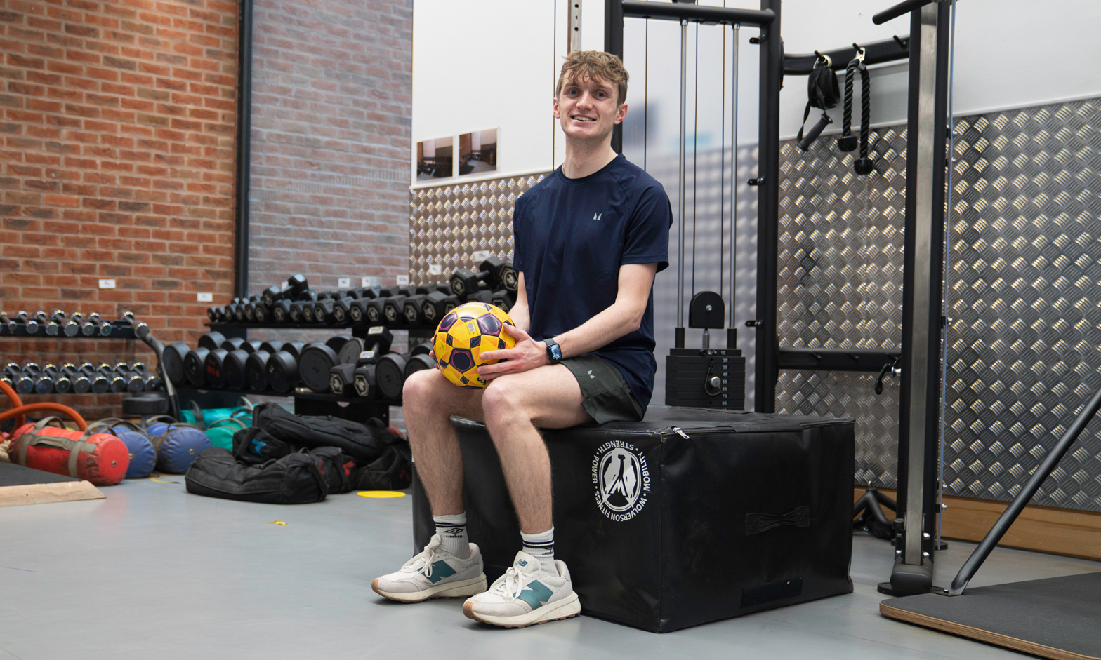 A male student sitting in a gym setting holding a football