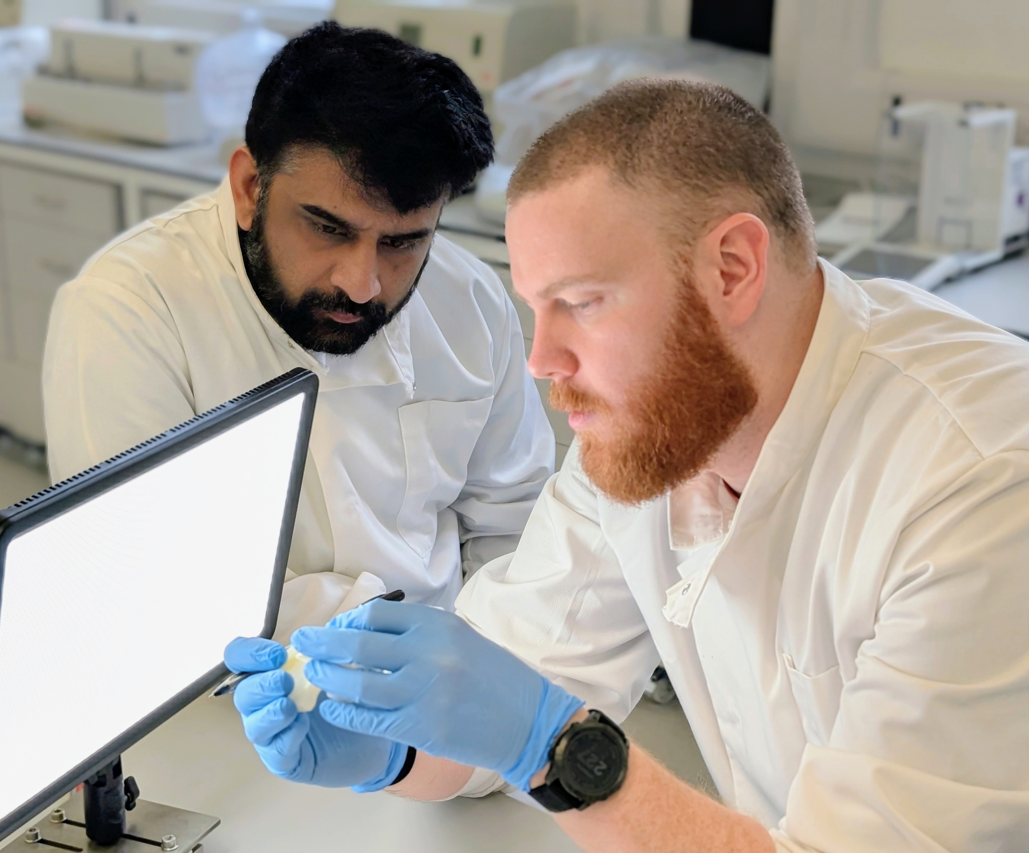 Two male scientists in while labcoats look closely at a small device and glowing screen.