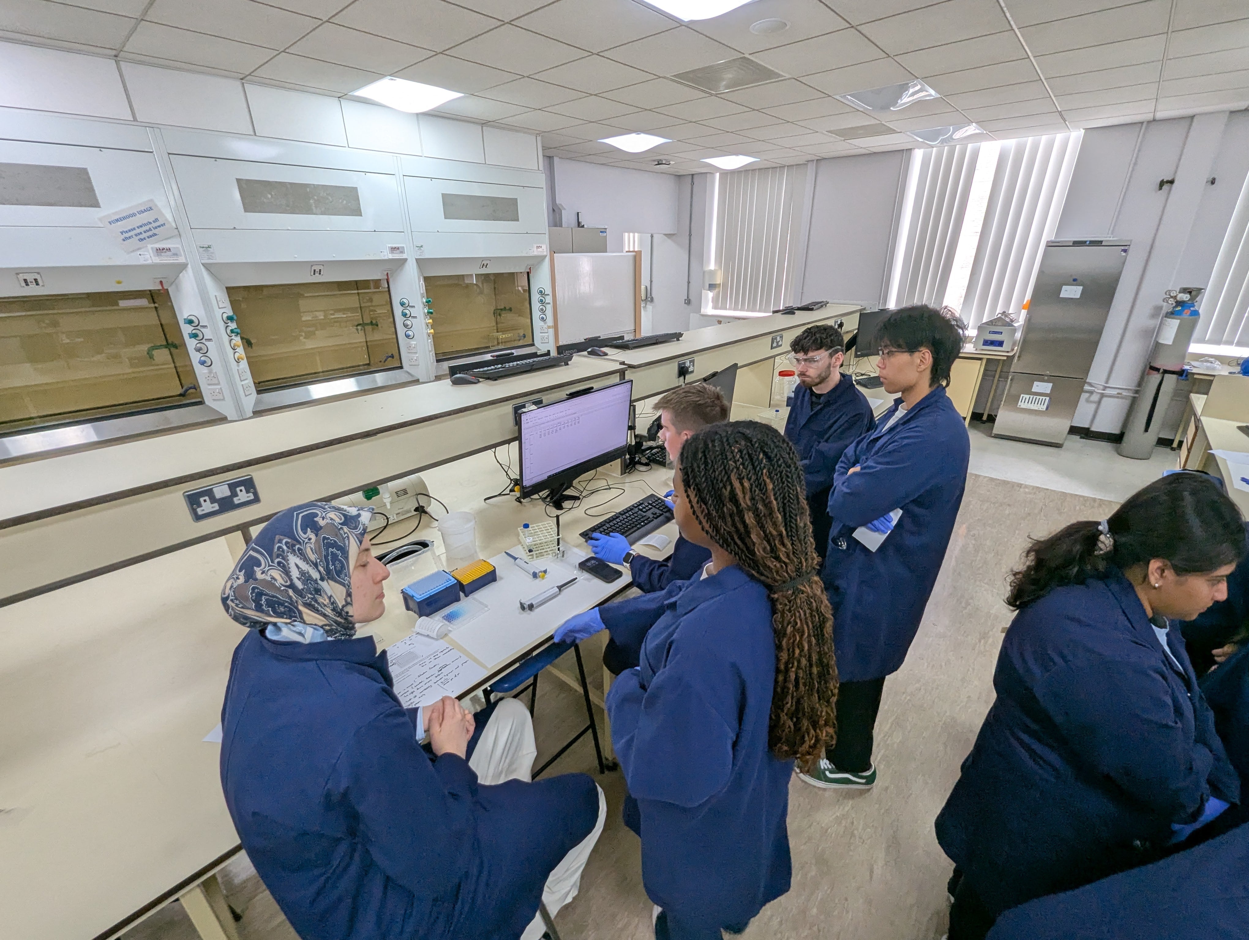 Summer school students in a sterile environment of a lab looking at the monitor and conducting an experiment