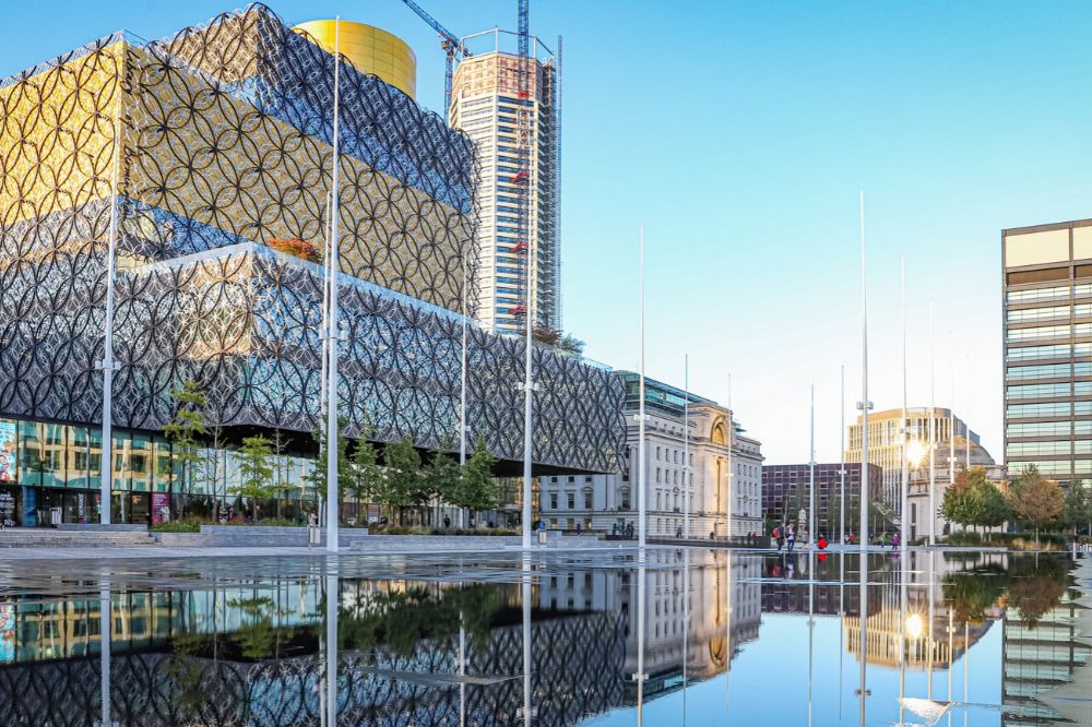 A large glass building with yellow accents and circular patterned architecture sits behind fountains in the foreground. 