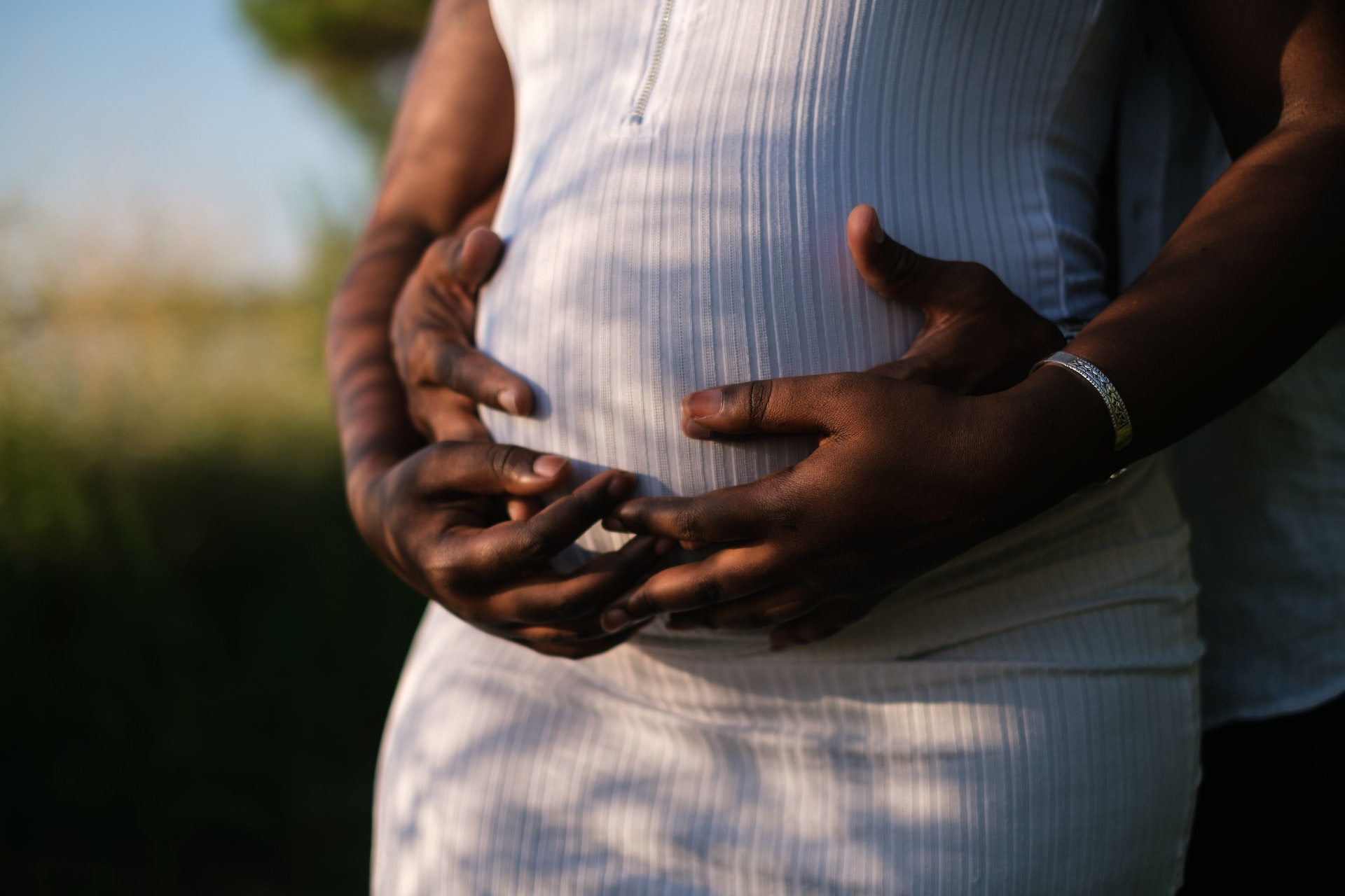 Black expectant mother wearing white dress with her hands and the hands of black father behind her on her pregnancy bump