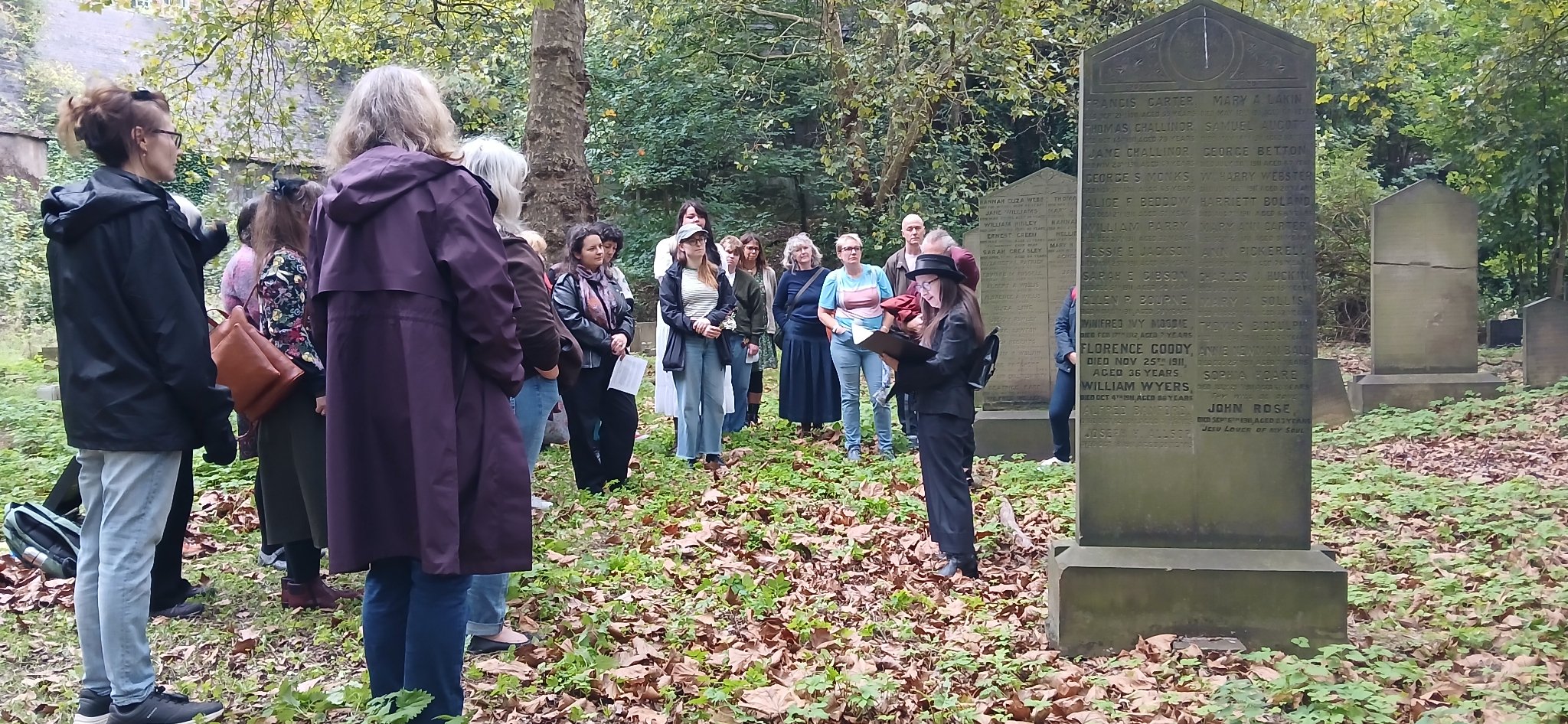 People gathered in an autumnal cemetery listening to a performer standing by a tall gravestone.