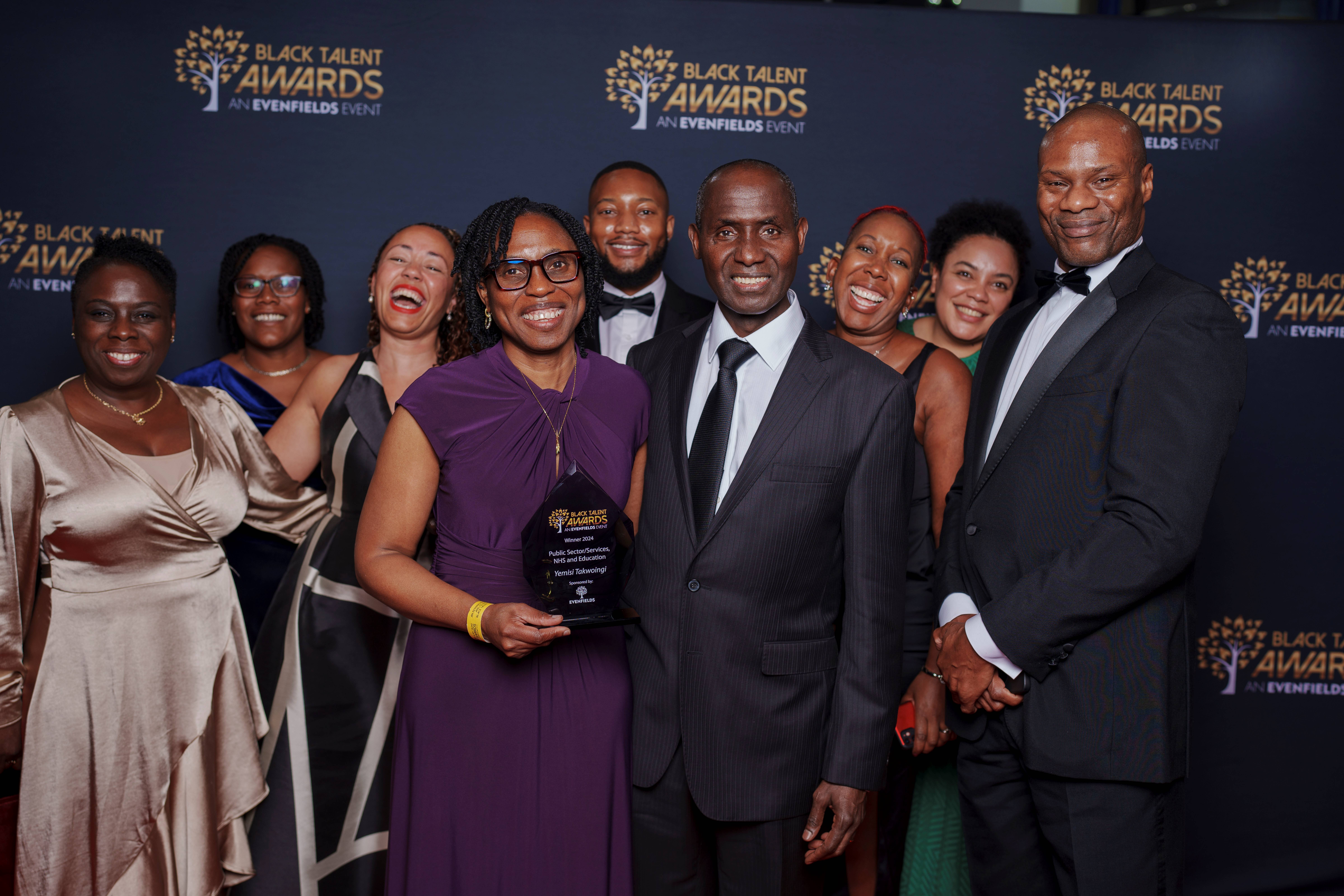 Yemisi holding her prize surrounded by a group of smiling people in 'black tie' clothing. 
