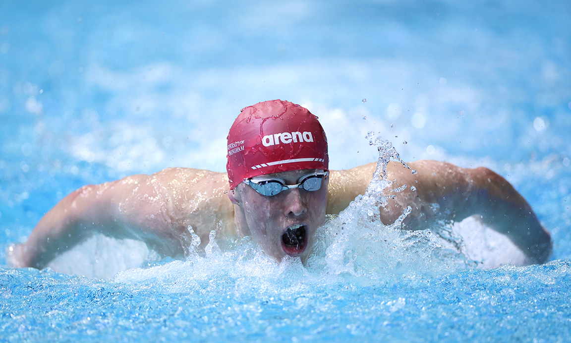 A University of Birmingham swimmer, facing the camera and swimming through the water with a swimming cap and goggles