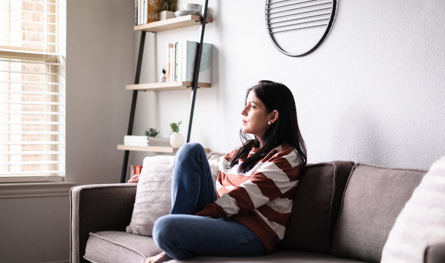A woman with brunette hair and striped top sits on a grey sofa, looking out of a window.