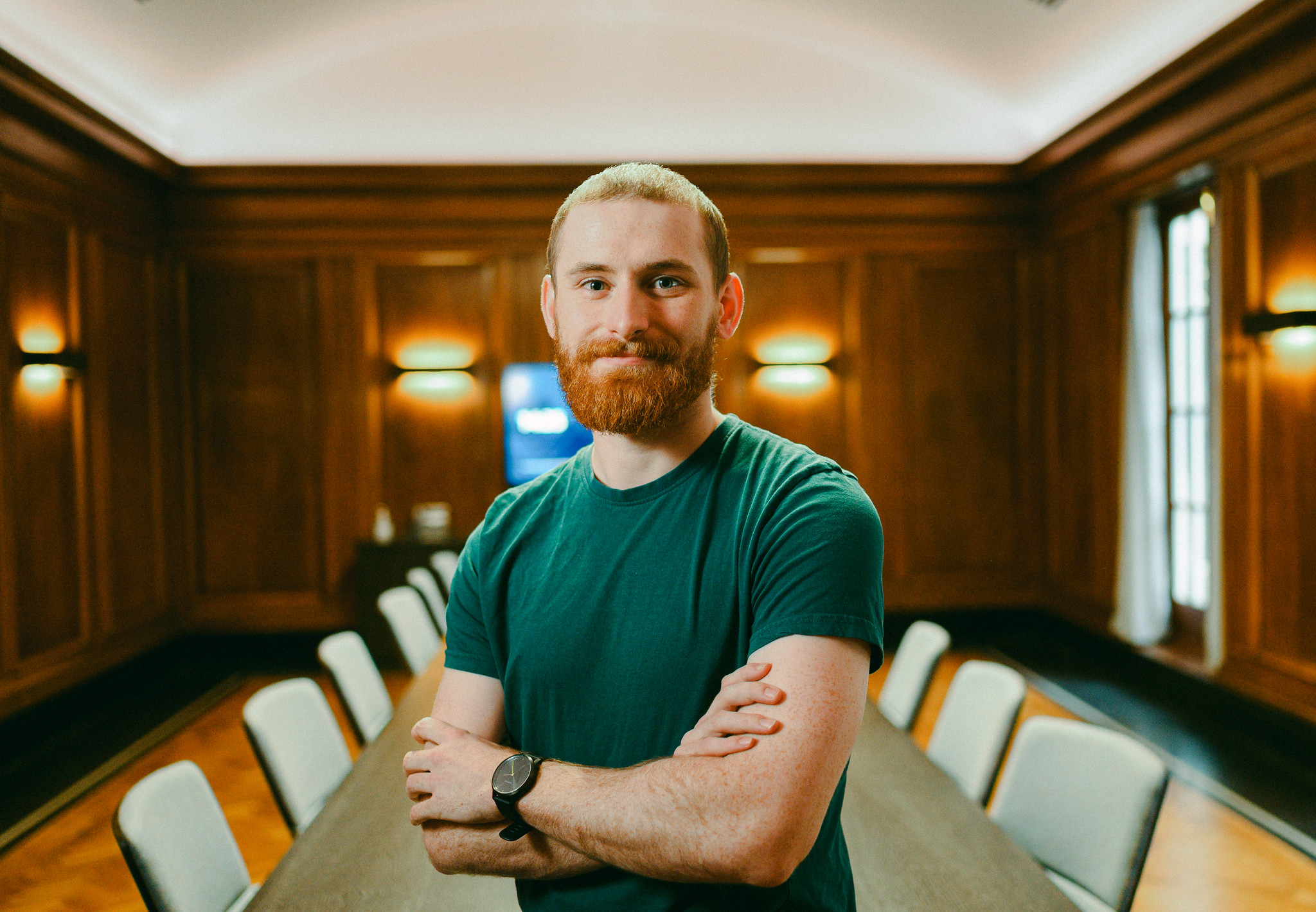John stood with arms folded in front of a table and chairs in a board room