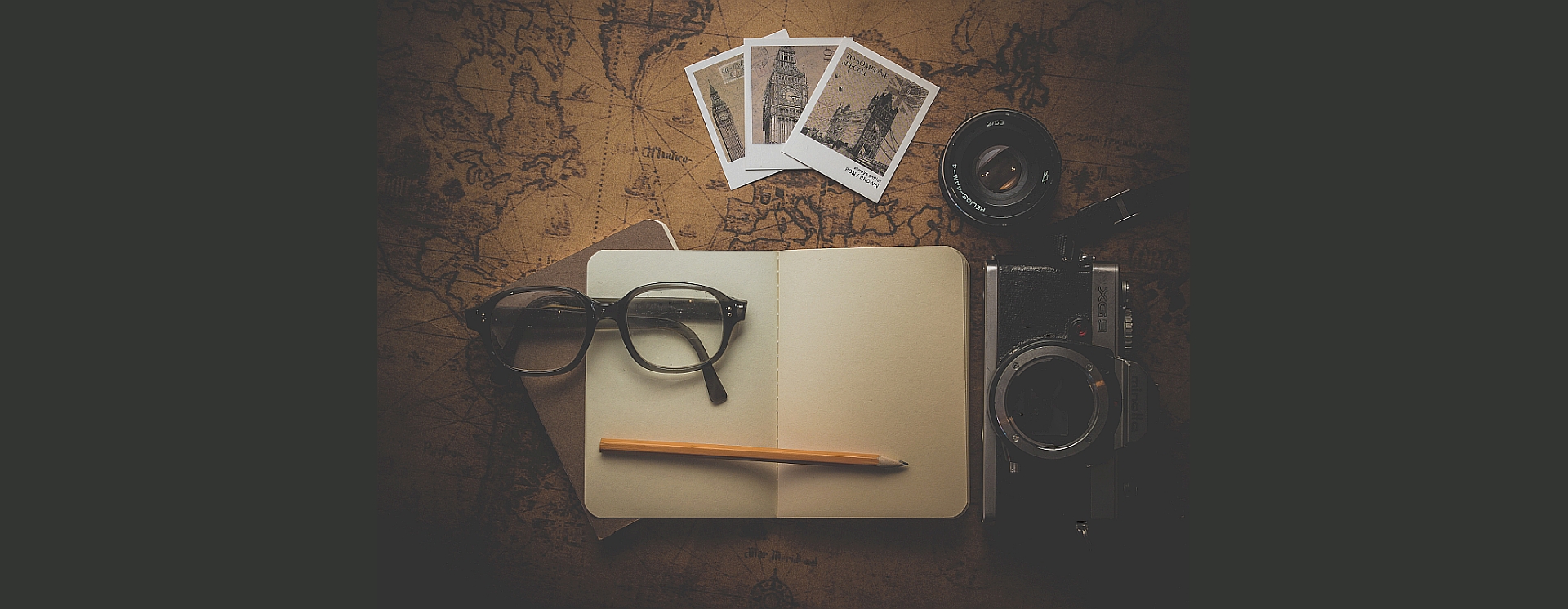 A pair of spectacles and a pencil rest on an open blank notebook. An old world map, camera and photographs are nearby.