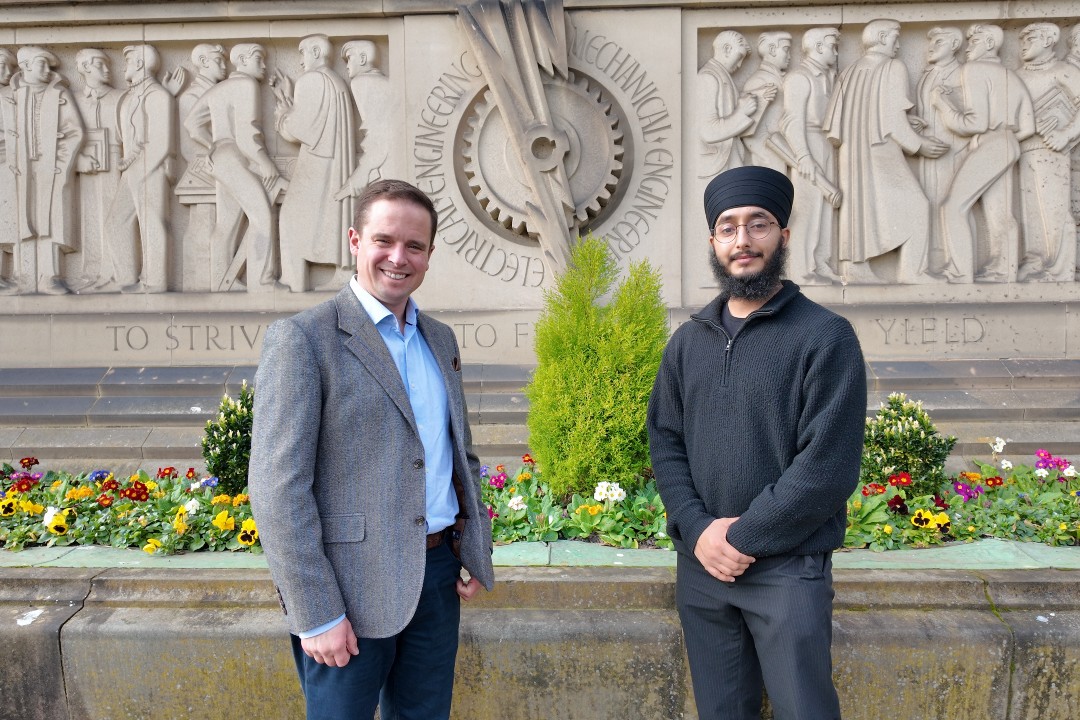 A man in a tweed jacket stands with a man in grey jumper and turban outside a frieze showing early engineers