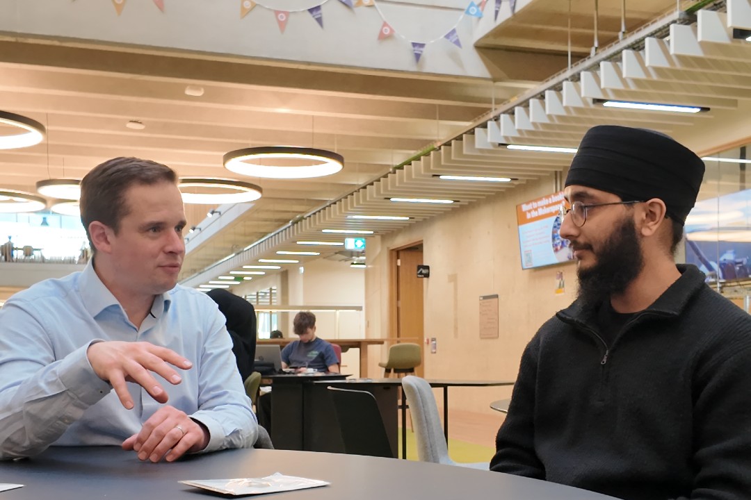 A man in open necked blue shirt sits at a table in discussion with man in grey jumper, glasses and turban
