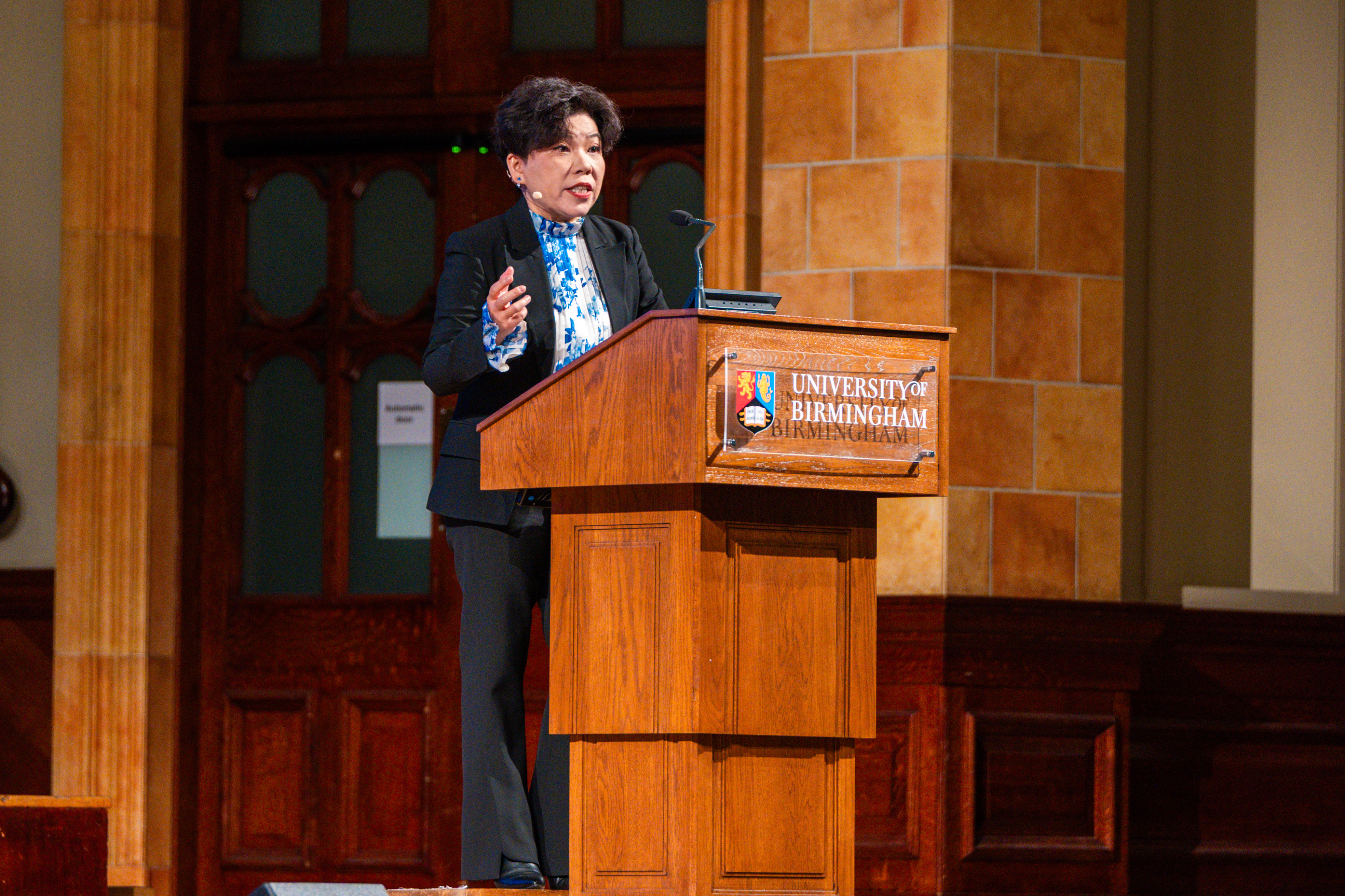 Dr Deng Yaping stands at a podium with the University of Birmingham logo attached to the front.