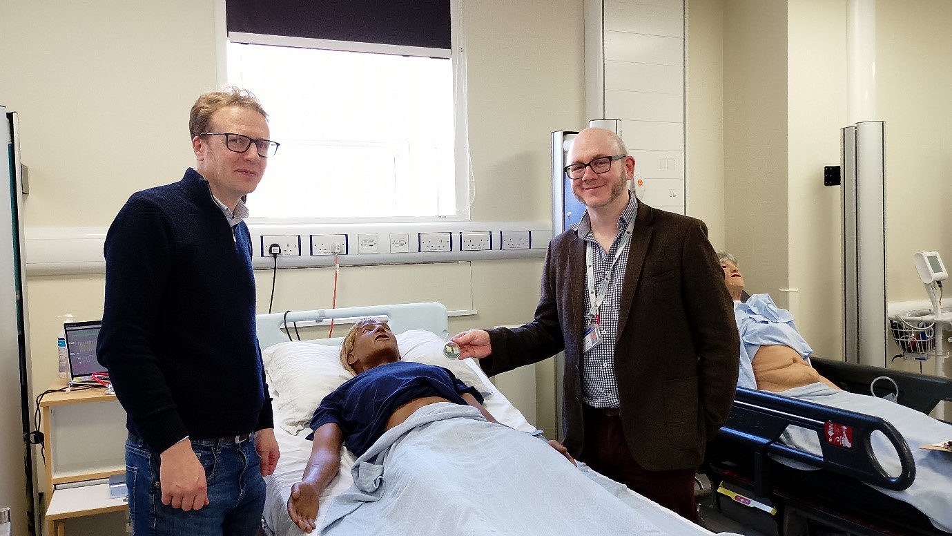 Two men stand either side of a hospital bed with a medical manikin in it. They are holding up the new medical device being tested.