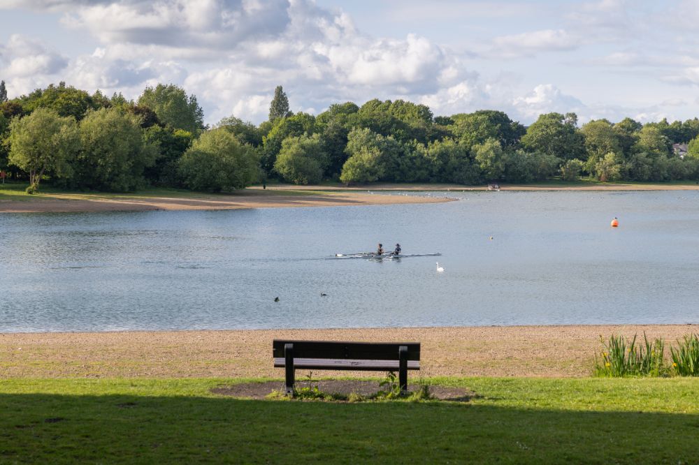 A reservoir of water surrounded by greenery and sandy shoreline and two people in boat