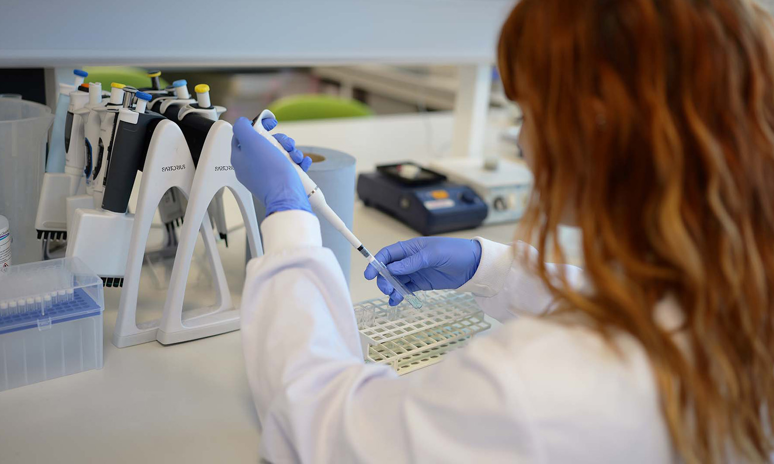 A female researcher wearing a white lab coat and purple Latex gloves dropping a solution into a test tube