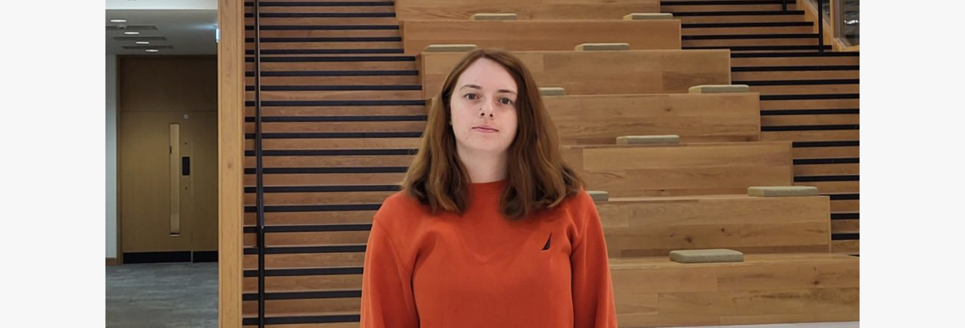 Caitlin, a female student with mid-length brown hair and an orange jumper, stands in front of a tall wooden staircase