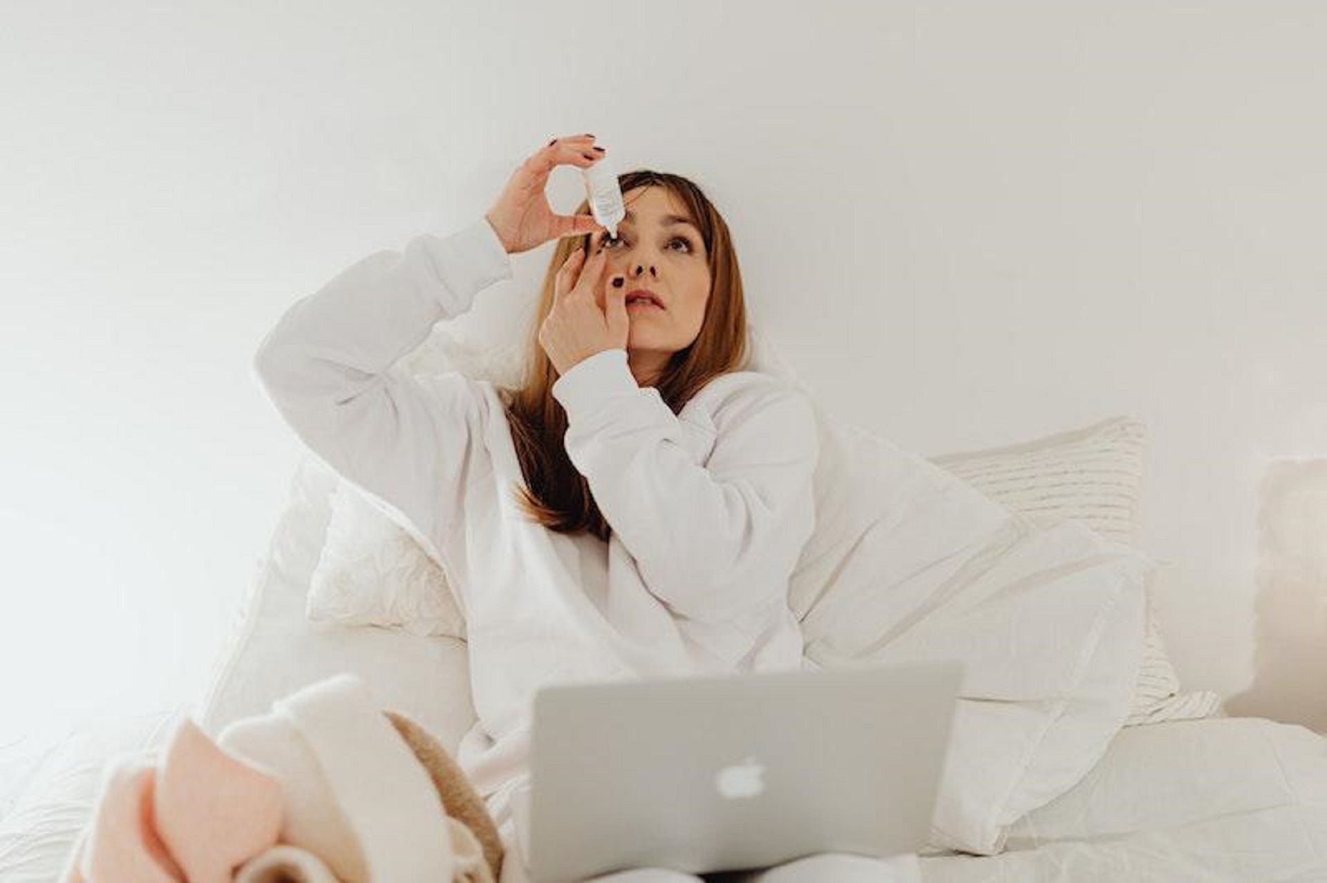    Woman with white top and auburn hair self-administering eye drop while sat on sofa with laptop   