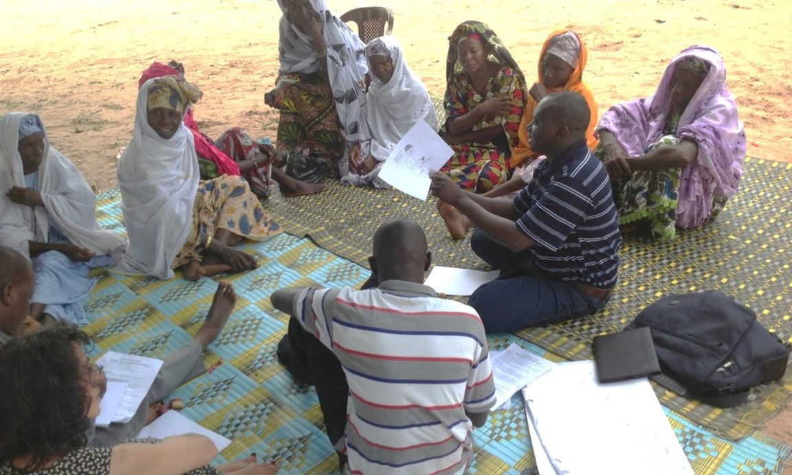 A group discussion between twelve people sat on mats on the ground outside in the Gambia.